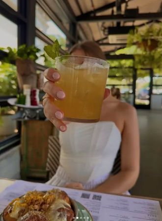 Hand holding an iced citrus cocktail with a mint garnish toward the camera at a sunlit, greenery-filled patio cafe with a pastry and menu on the table — casual brunch vibe.