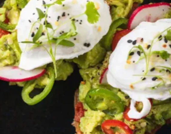 Close-up of avocado toast with smashed avocado, two poached eggs, sliced radishes, jalapeño rings, microgreens and black sesame seeds — bright zesty brunch dish