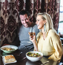 Smiling couple at a cozy restaurant table on a casual date night, woman holding a glass of white wine with bowls of salad and a piece of bread on the table against a patterned curtain backdrop
