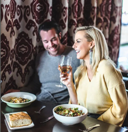 Smiling couple at a cozy restaurant table on a casual date night, woman holding a glass of white wine with bowls of salad and a piece of bread on the table against a patterned curtain backdrop