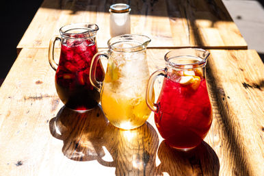 Three glass pitchers of iced summer drinks—two red fruit punch/sangria-style and one golden iced tea or lemonade with lemon slices—on a sunlit wooden table casting bright shadows.