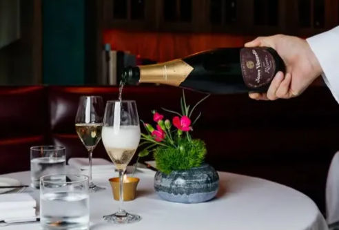 Hand pouring champagne into a flute at an upscale downtown restaurant table with white tablecloth, water glasses, lit votive and small pink floral centerpiece — elegant romantic fine-dining scene.