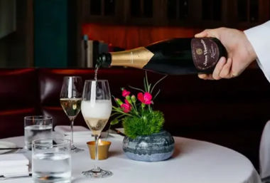 Hand pouring champagne into a flute at an upscale downtown restaurant table with white tablecloth, water glasses, lit votive and small pink floral centerpiece — elegant romantic fine-dining scene.
