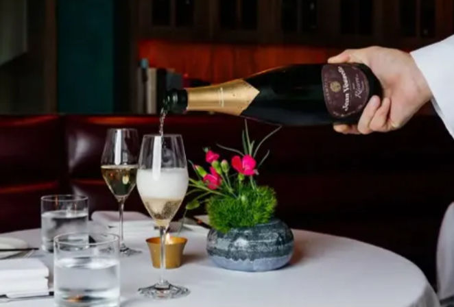 Hand pouring champagne into a flute at an upscale downtown restaurant table with white tablecloth, water glasses, lit votive and small pink floral centerpiece — elegant romantic fine-dining scene.