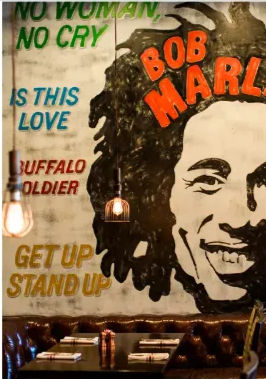Vibrant reggae mural of a smiling dreadlocked musician with song titles like 'No Woman No Cry', 'Is This Love', and 'Buffalo Soldier' painted on a brick restaurant wall above cozy leather booths and glowing pendant lights.