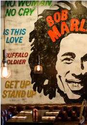 Vibrant reggae mural of a smiling dreadlocked musician with song titles like 'No Woman No Cry', 'Is This Love', and 'Buffalo Soldier' painted on a brick restaurant wall above cozy leather booths and glowing pendant lights.
