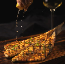 Hand squeezing lemon over Mediterranean-style grilled flatbread with herbs and char marks on a wooden board, glass of white wine blurred in the background