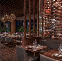 Warm-lit upscale restaurant interior with wooden tables set for dining, quilted chairs, thick rope dividers, curving wood ceiling, globe pendant lights and a glass-enclosed wine rack wall.