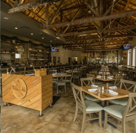 Rustic modern restaurant interior with exposed wooden beams and vaulted ceiling, long bar, rows of wooden tables set with white napkins and glassware on a polished concrete floor, cozy farmhouse-style dining space.