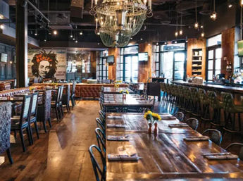 Rustic-industrial restaurant interior with long communal wooden tables, leather banquette, chandelier-lit bar seating, exposed brick, windows and small yellow flower centerpieces