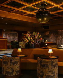 Warm, retro upscale hotel lounge with amber lighting: plush curved banquette seating, patterned barrel chairs with tiger motifs, small round tables with glowing lamps, a large floral centerpiece and a mirrored disco ball under a geometric coffered ceiling.