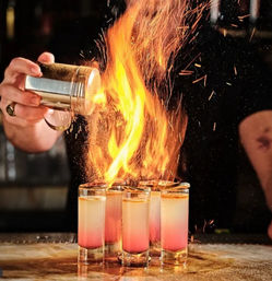 Flaming cocktail shots at a bar: bartender sprinkling garnish over a row of pink-to-clear layered shot glasses as bright orange flames and sparks leap up.
