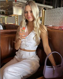 Smiling woman in white crop top and linen pants sipping a pink cocktail garnished with watermelon in a modern restaurant booth, lavender handbag beside her.