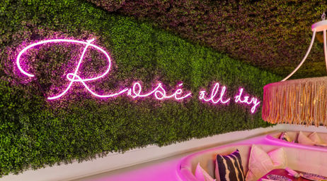 Chic indoor lounge corner with pink "Rosé all day" neon sign on a green plant wall, curved banquette seating and a fringed pendant lamp