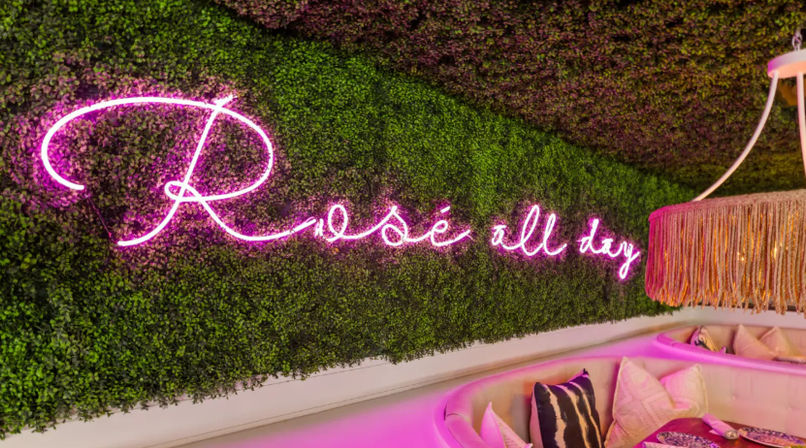 Chic indoor lounge corner with pink "Rosé all day" neon sign on a green plant wall, curved banquette seating and a fringed pendant lamp