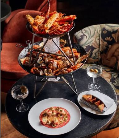 Two-tier seafood tower piled with crab legs, clams and shrimp on a round bistro table, paired with seared scallops on a red purée, toasted bread triangles and two martini glasses in an elegant seafood restaurant setting.