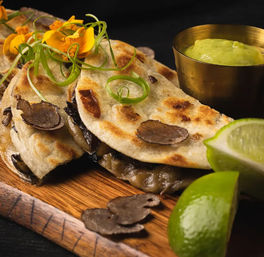 Gourmet truffle and mushroom quesadillas on charred flatbreads with zesty lime wedges, green avocado crema in a brass cup, scallion curls and edible flowers on a wooden board.