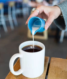Hand pouring cream from a small blue pitcher into black coffee in a white mug on a wooden cafe table