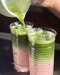 Close-up of two layered iced matcha lattes: bright green matcha being poured from a pitcher over pastel pink milk and ice in tall ribbed glasses.
