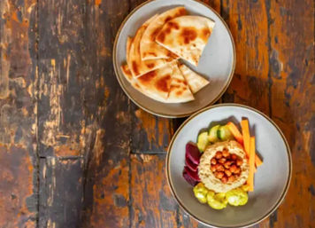 Top-down view of a Mediterranean hummus bowl topped with roasted chickpeas, pickled beets, cucumber rounds and carrot sticks, with sliced pita on a rustic wooden table.
