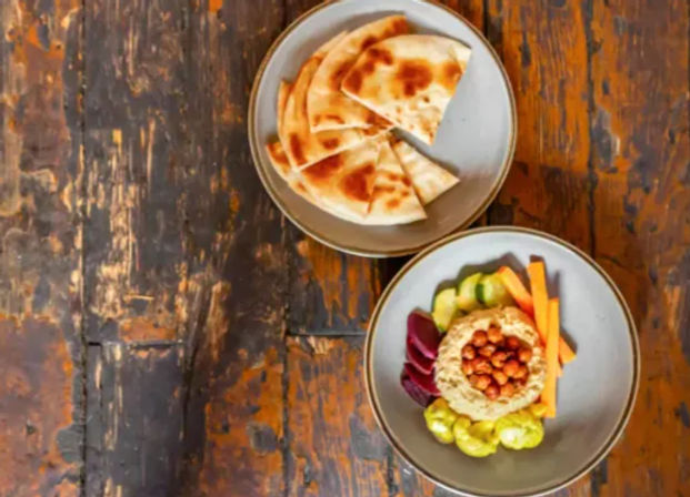 Top-down view of a Mediterranean hummus bowl topped with roasted chickpeas, pickled beets, cucumber rounds and carrot sticks, with sliced pita on a rustic wooden table.