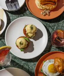 Top-down brunch flat lay: two hollandaise-topped eggs benedict on a white plate, pancakes and a fried-egg sandwich on orange plates, and citrus cocktails on a textured green tablecloth.