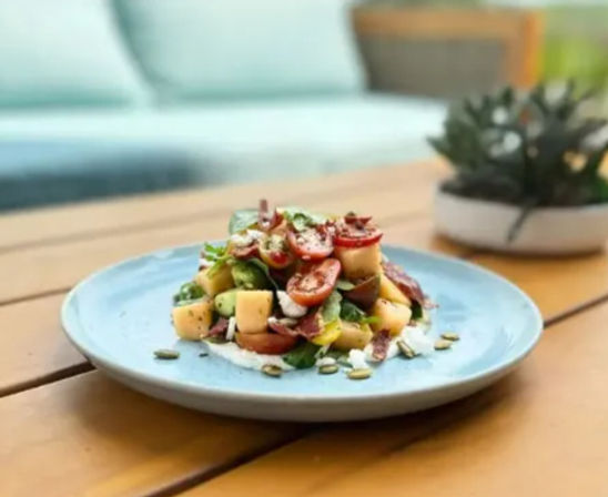 Bright summer salad of cherry tomatoes, cucumber, cheese cubes, red onion and pumpkin seeds on a blue plate on a wooden outdoor patio table with blurred sofa and succulent in background
