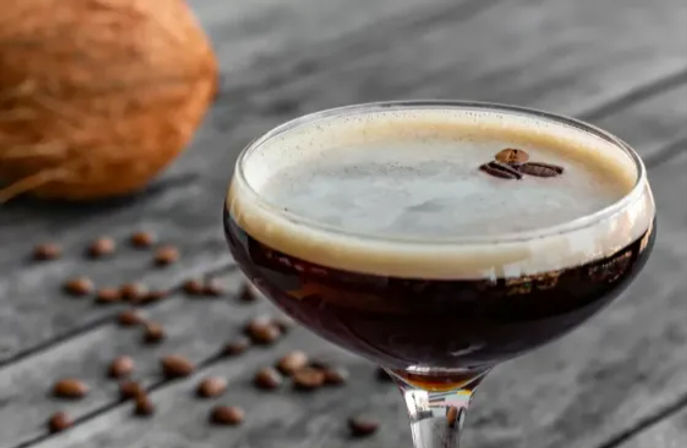 Espresso martini in a coupe glass with creamy foam and three coffee beans on top, scattered coffee beans and a blurred coconut on a rustic wooden table.