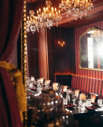 Intimate upscale restaurant dining room with a long set table, burgundy velvet banquette seating, crystal chandeliers, gold-framed mirror and candlelit glassware — elegant fine-dining interior.