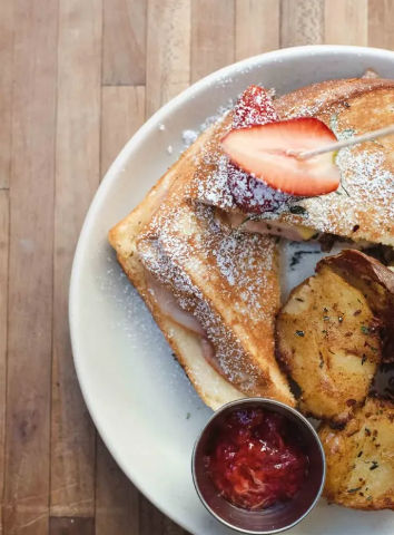 Overhead shot of golden French toast dusted with powdered sugar and garnished with a sliced strawberry, served with roasted herb potatoes and a ramekin of strawberry jam on a white plate over a wooden tabletop.