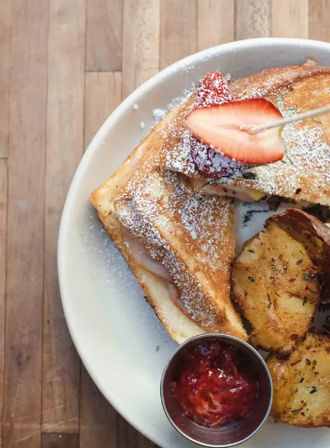 Overhead shot of golden French toast dusted with powdered sugar and garnished with a sliced strawberry, served with roasted herb potatoes and a ramekin of strawberry jam on a white plate over a wooden tabletop.