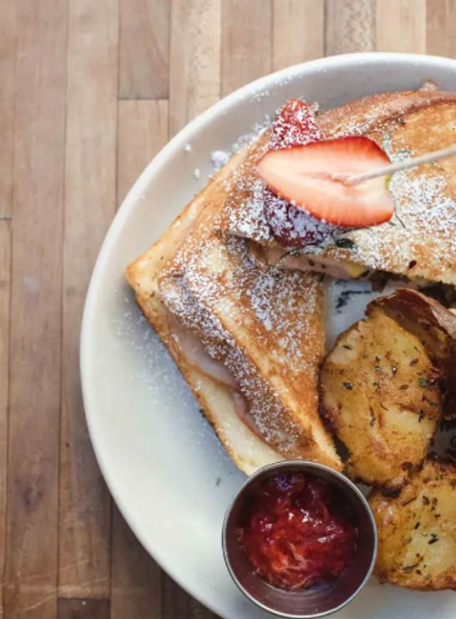 Overhead shot of golden French toast dusted with powdered sugar and garnished with a sliced strawberry, served with roasted herb potatoes and a ramekin of strawberry jam on a white plate over a wooden tabletop.