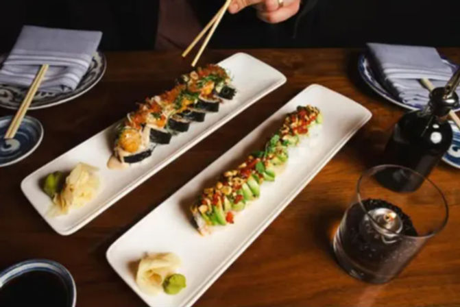 Two long rectangular plates of sushi rolls on a wooden restaurant table — one topped with avocado and crunchy bits, the other with spicy mayo and nori; chopsticks, soy sauce, pickled ginger and wasabi nearby.
