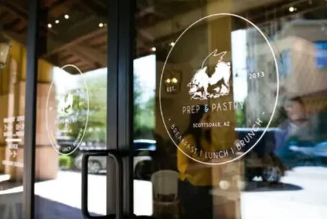 Glass cafe entrance with a circular decal featuring a winged-pig logo, reflections of cars and people outside, and black door pull handles.