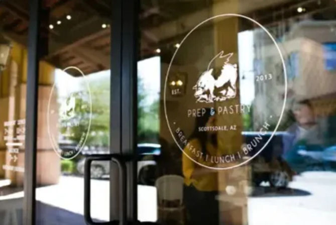 Glass cafe entrance with a circular decal featuring a winged-pig logo, reflections of cars and people outside, and black door pull handles.