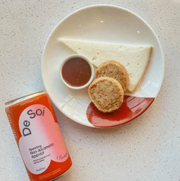 White plate with a triangular wedge of cheese, two round crackers and a small ramekin of brown dipping sauce, paired with an orange can of sparkling non-alcoholic aperitif on a speckled countertop — a light snack pairing.
