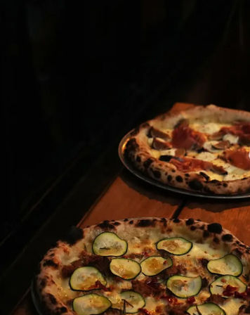 Two Neapolitan-style wood-fired pizzas on metal trays on a rustic wooden table in a dimly lit pizzeria — front pizza topped with zucchini slices and melted cheese, back pizza with cured meat.