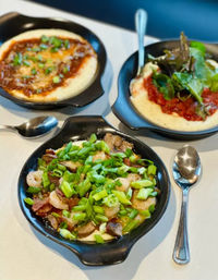 Three black skillets of creamy grits for Southern-style brunch: front bowl with shrimp, bacon, mushrooms and chopped green onions; two bowls in background with tomato topping, mixed greens and melted cheese, served with spoons.