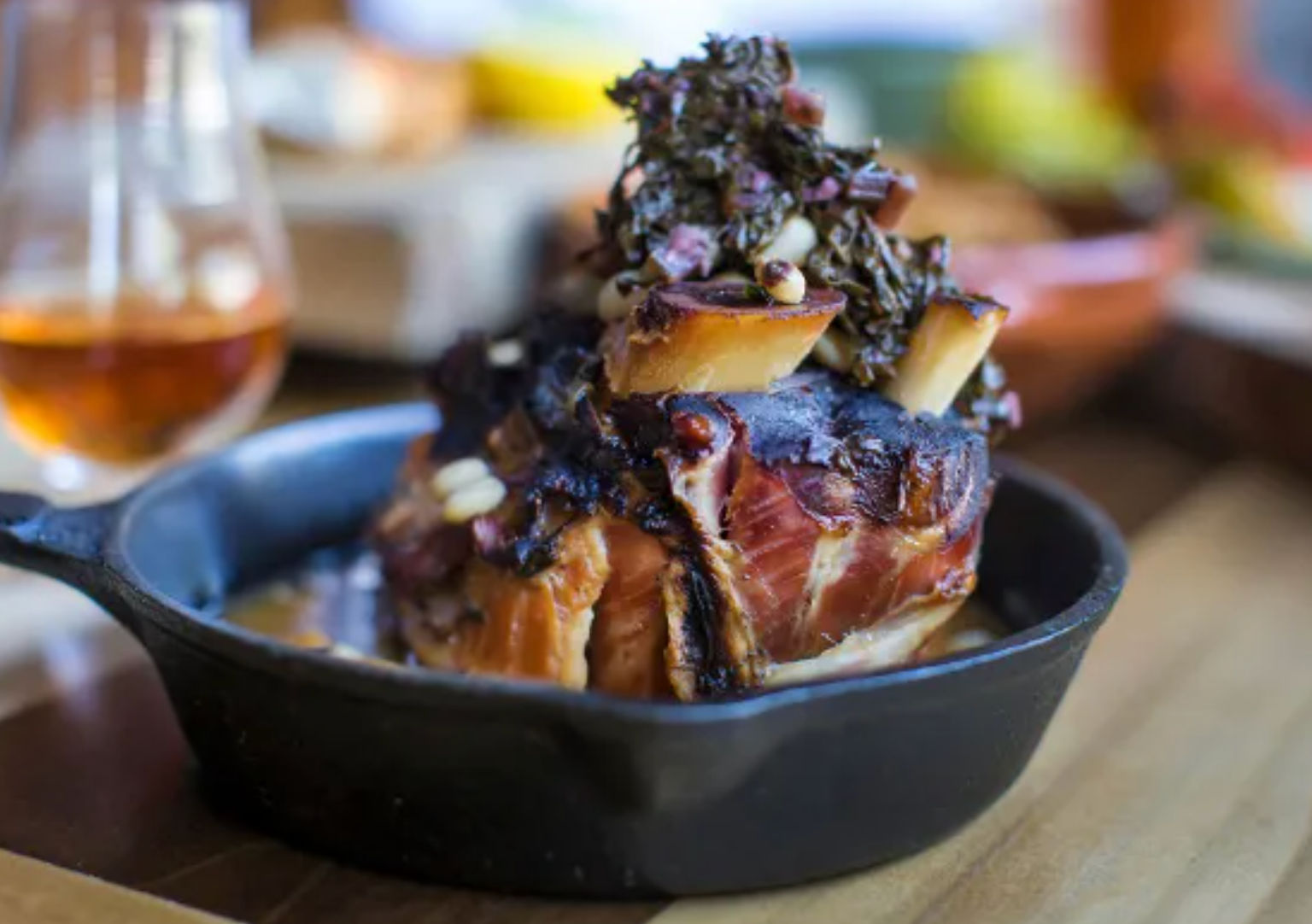 Gourmet braised beef shank served in a cast-iron skillet, topped with roasted bone marrow, chopped herbs and rich jus, with a blurred glass of amber drink in the background.