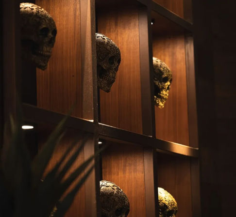 Ornate decorative skulls peeking from wood-paneled cubby shelves under warm, moody lighting — spooky-chic interior shelf display for bar or lounge decor