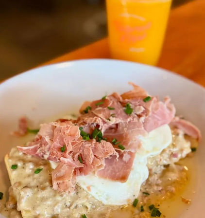 Brunch plate of poached eggs and shaved ham atop creamy grits, sprinkled with chopped chives; glass of orange juice blurred in the background.