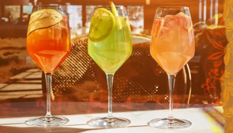 Three bright, refreshing spritz cocktails — orange, green, and pink — garnished with citrus slices and served in wine glasses on a sunlit restaurant patio table.