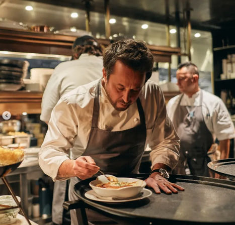 Chef in an open-kitchen restaurant carefully garnishing a bowl of soup on the pass, wearing a white jacket and apron during service.