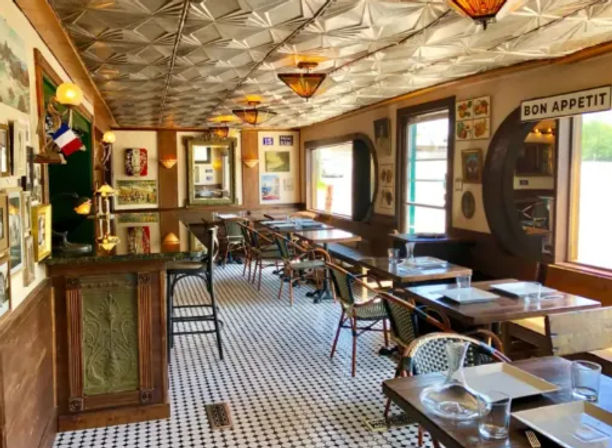Cozy French-inspired bistro interior with pressed-tin ceiling, black-and-white mosaic tile floor, rattan café chairs, small tables set by large windows and a “Bon Appetit” wall sign.