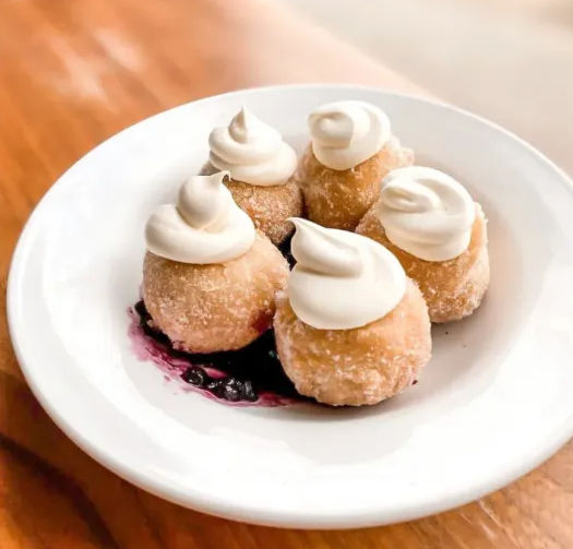 Five bite-sized sugared doughnut holes crowned with swirls of whipped cream, arranged on a white plate over a pool of blueberry compote on a wooden table.