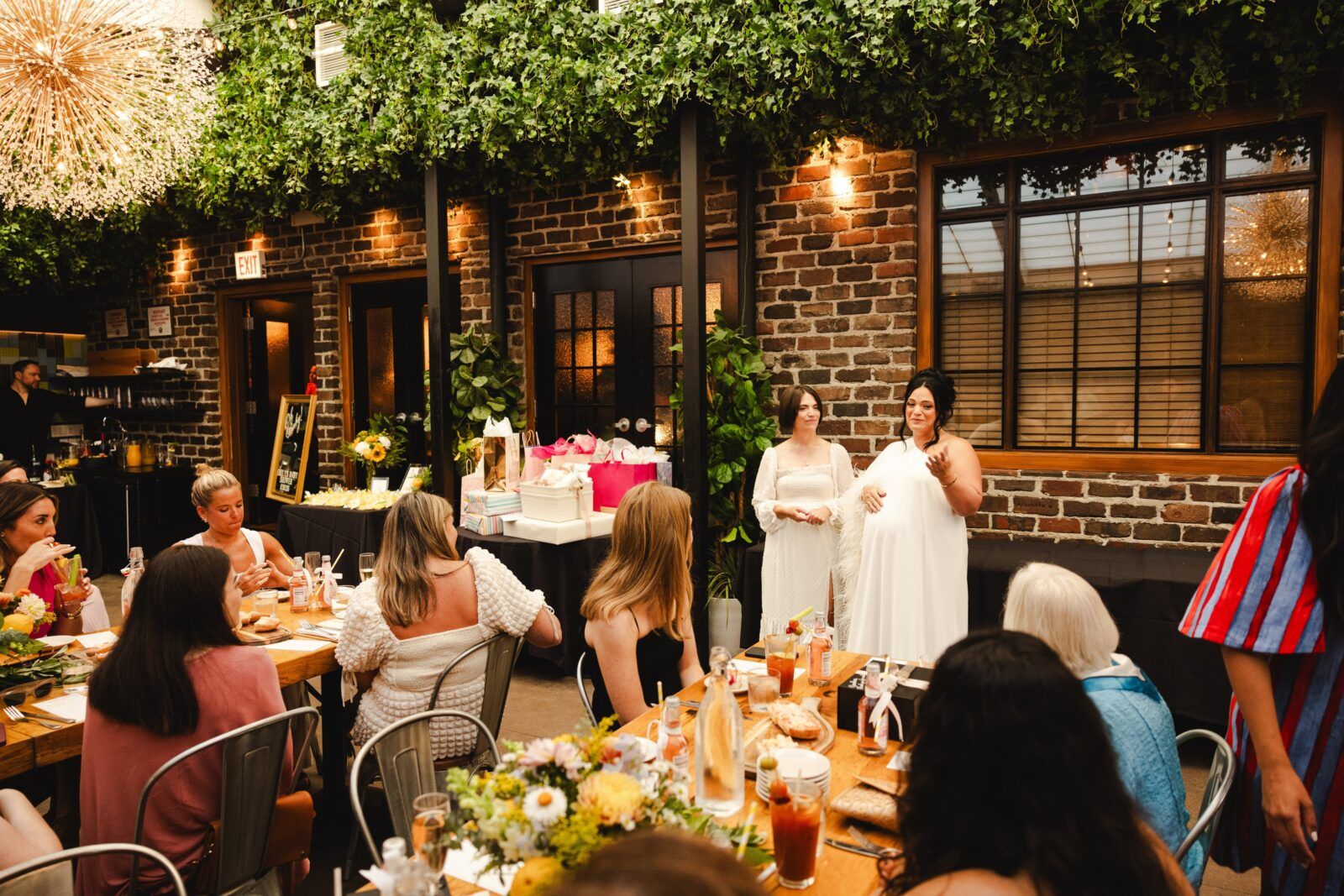 Mom-to-be speaking at a cozy indoor garden-style baby shower in a brick-walled venue, guests seated at a long wooden brunch table with floral centerpieces, drinks, and a table piled with wrapped gifts.