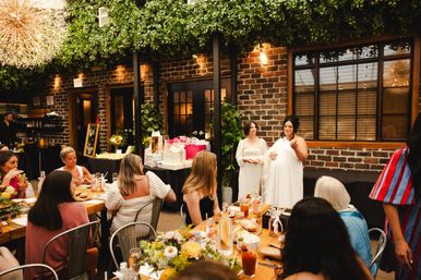 Mom-to-be speaking at a cozy indoor garden-style baby shower in a brick-walled venue, guests seated at a long wooden brunch table with floral centerpieces, drinks, and a table piled with wrapped gifts.