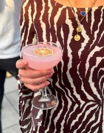Hand holding a pink cocktail in a coupe glass topped with a dried citrus slice, set against a zebra-print top and layered necklaces.