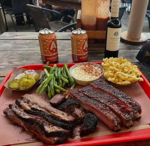 Mouthwatering smoked barbecue platter on a red tray on a rustic outdoor table: sliced brisket and beef ribs with dark bark, creamy mac and cheese, green beans, pickles and onions, a small creamy side, and two beer cans plus a bottle of wine in the background.