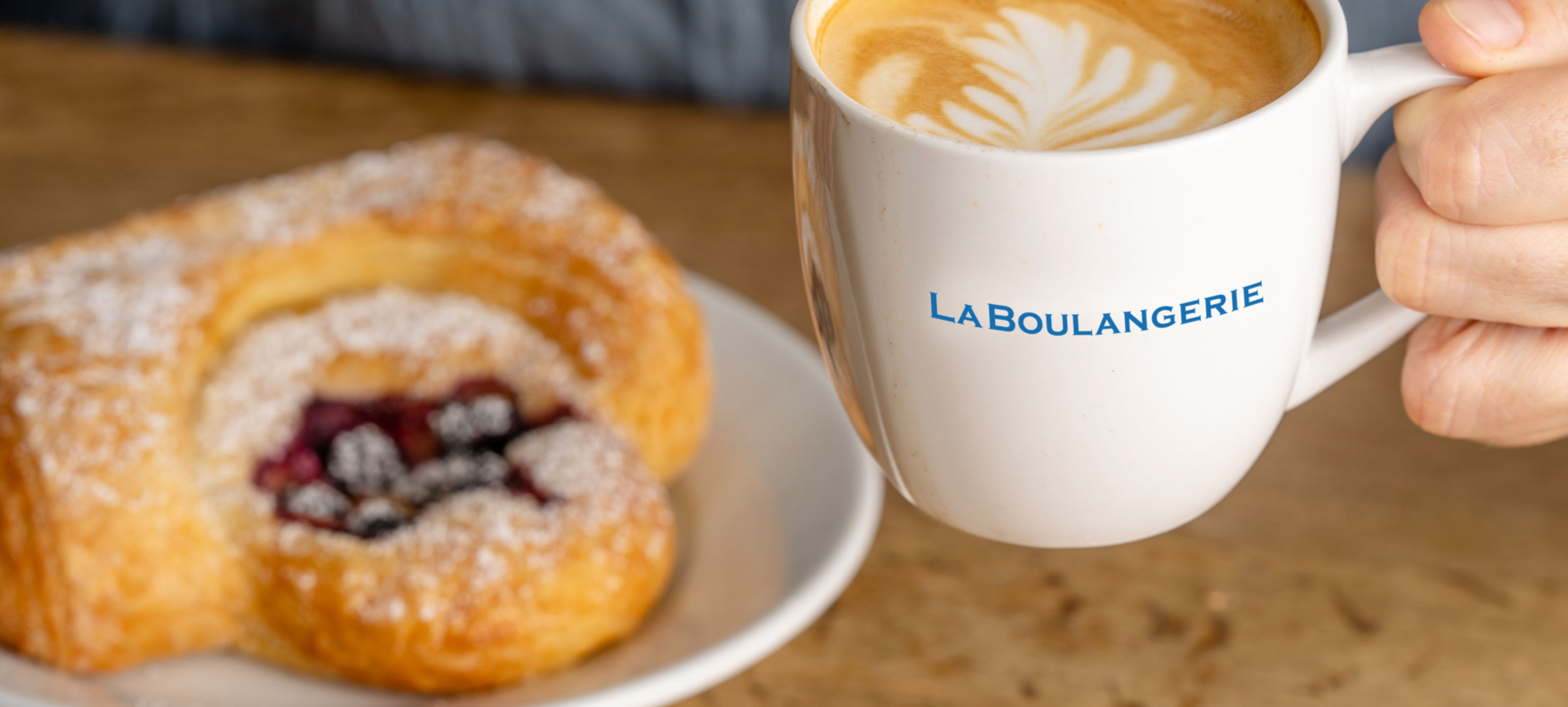 Hand holding a white ceramic mug of latte with leaf latte art beside a powdered-sugar berry Danish on a white plate, set on a wooden café table.